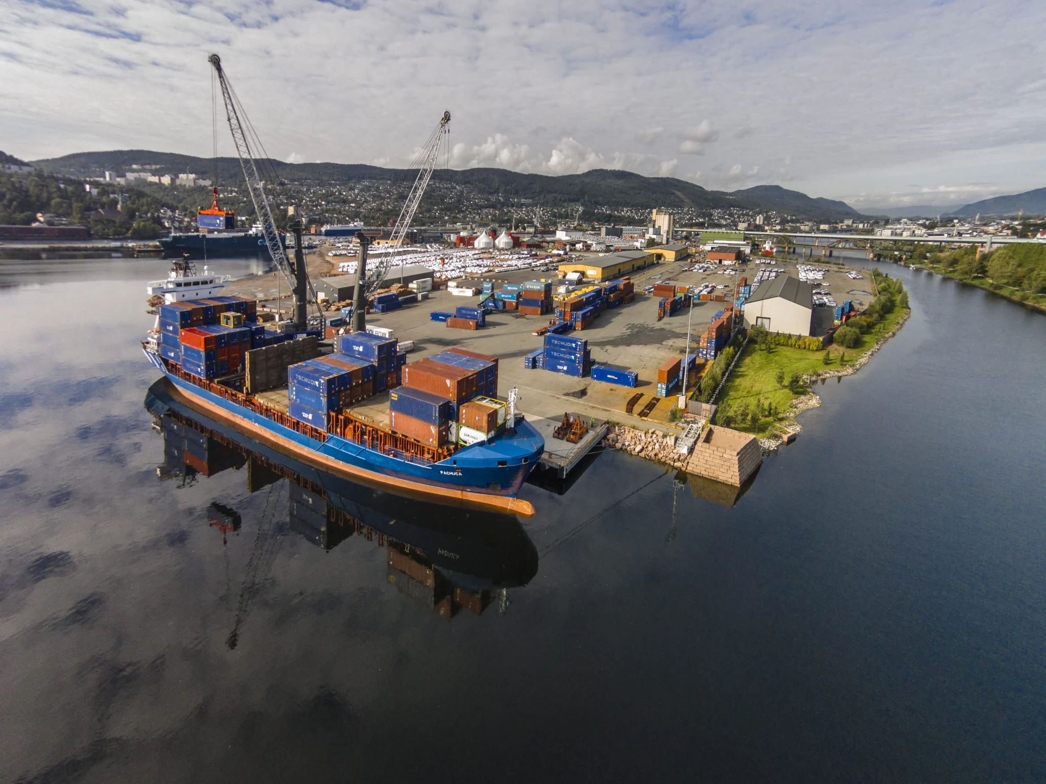 A large container ship docked at a busy port terminal with cranes loading and unloading cargo.